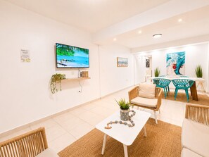 A modern living room with a TV, a dining area with chairs, and a kitchen in the background.