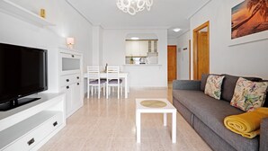 Una sala moderna con una televisión de pantalla plana, un sofá, una mesa de centro, un comedor con sillas y una cocina al fondo.