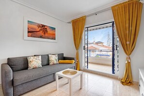 A living room with a sofa, a small table, a framed picture, and a view through a glass door. - Apartment 'Parque Mar V' with Shared Pool, Wi-Fi and Air Conditioning (La Mata)