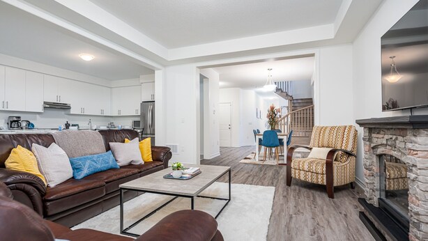 A modern living room with a brown leather sofa, a coffee table, and a fireplace. There is a kitchen in the background with white cabinets and a dining area with a blue chair. - Luxury Villa 5BR-4.5 Bath (Springwater)