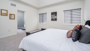 A bedroom with a large bed, two windows with blinds, a door leading to a bathroom, and a small seating area. - Luxury Villa 5BR-4.5 Bath (Springwater)