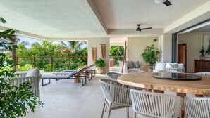 Un vaste espace extérieur doté d’une table ronde en bois et de chaises en osier blanches, entouré de fauteuils et de plantes en pot.