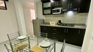 Cozinha compacta com armários escuros, micro-ondas e fogão a gás. Há uma mesa de vidro com cadeiras e uma porta branca ao fundo.