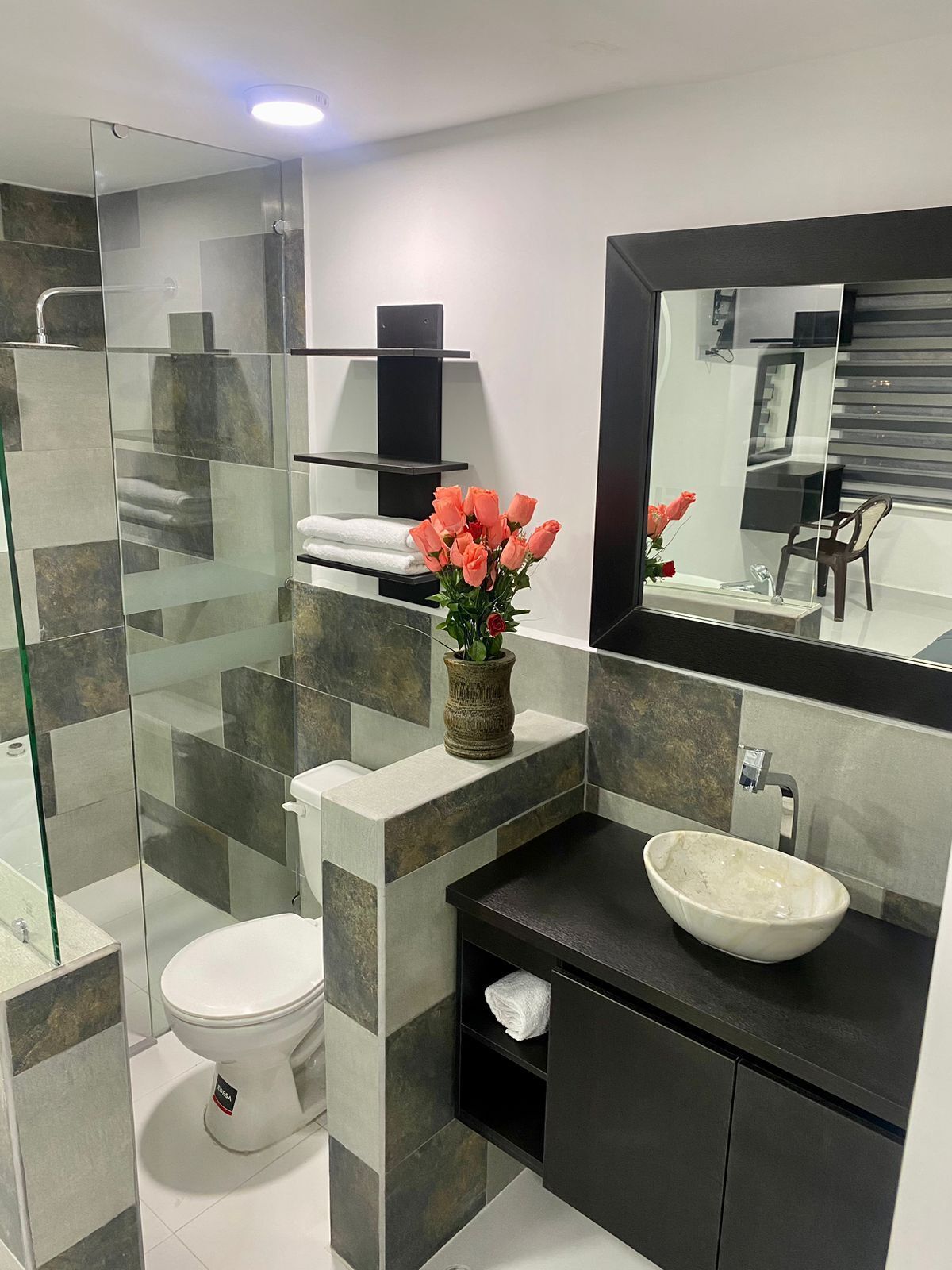 Une salle de bain moderne avec une douche à l’italienne en verre, des toilettes blanches, un meuble-lavabo noir avec un grand miroir et un vase rempli de fleurs.