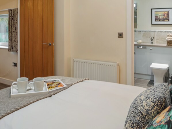 A hotel room with a bed, a tray with cups and a magazine, a wooden door, a radiator, and a bathroom visible in the background.