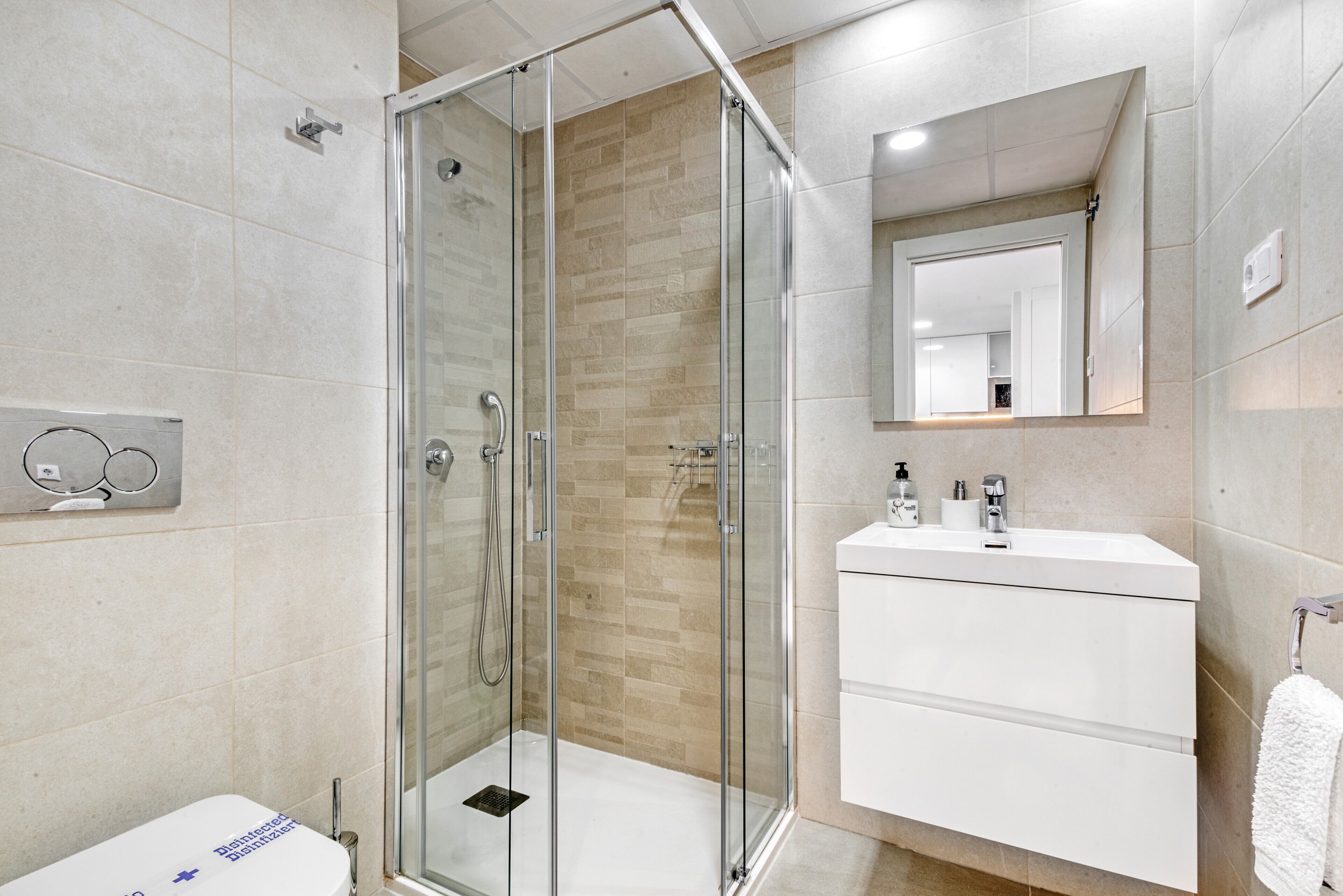 Un baño moderno con ducha de vidrio, lavamanos blanco y espejo sobre el lavamanos.