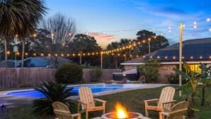 Un patio trasero con piscina, sillas de madera, un hogar para fuego y luces colgantes.