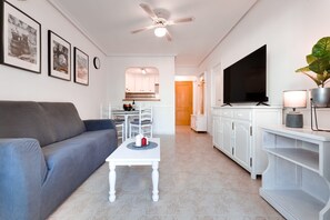 A living room with a grey sofa, a white coffee table, a flat-screen TV, a ceiling fan, and framed pictures on the wall.