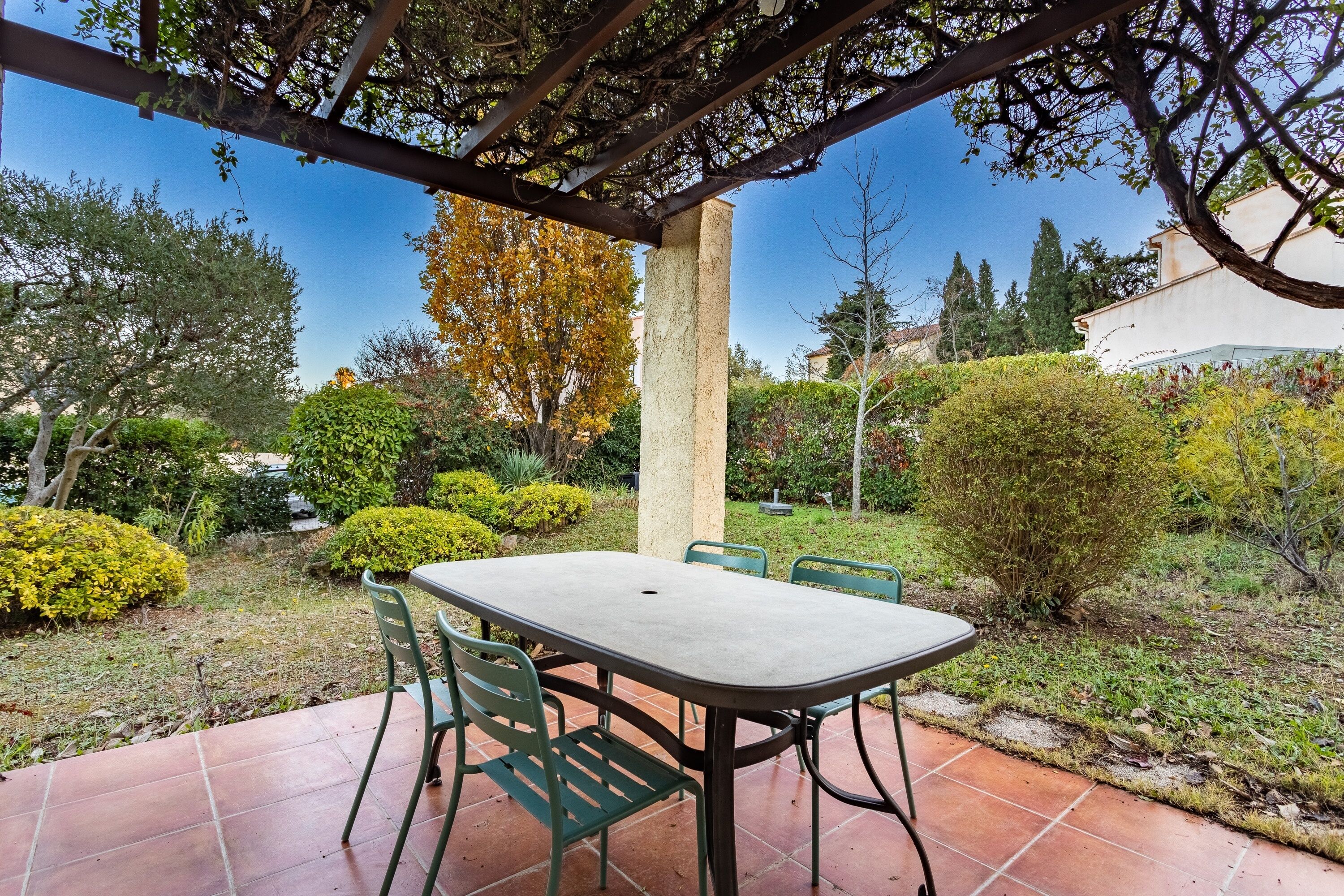 A covered patio with a table and chairs, surrounded by greenery and a clear sky.