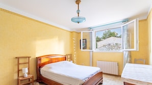A bedroom with a wooden bed, a bedside table with a lamp, a window with a view of greenery, and a radiator.