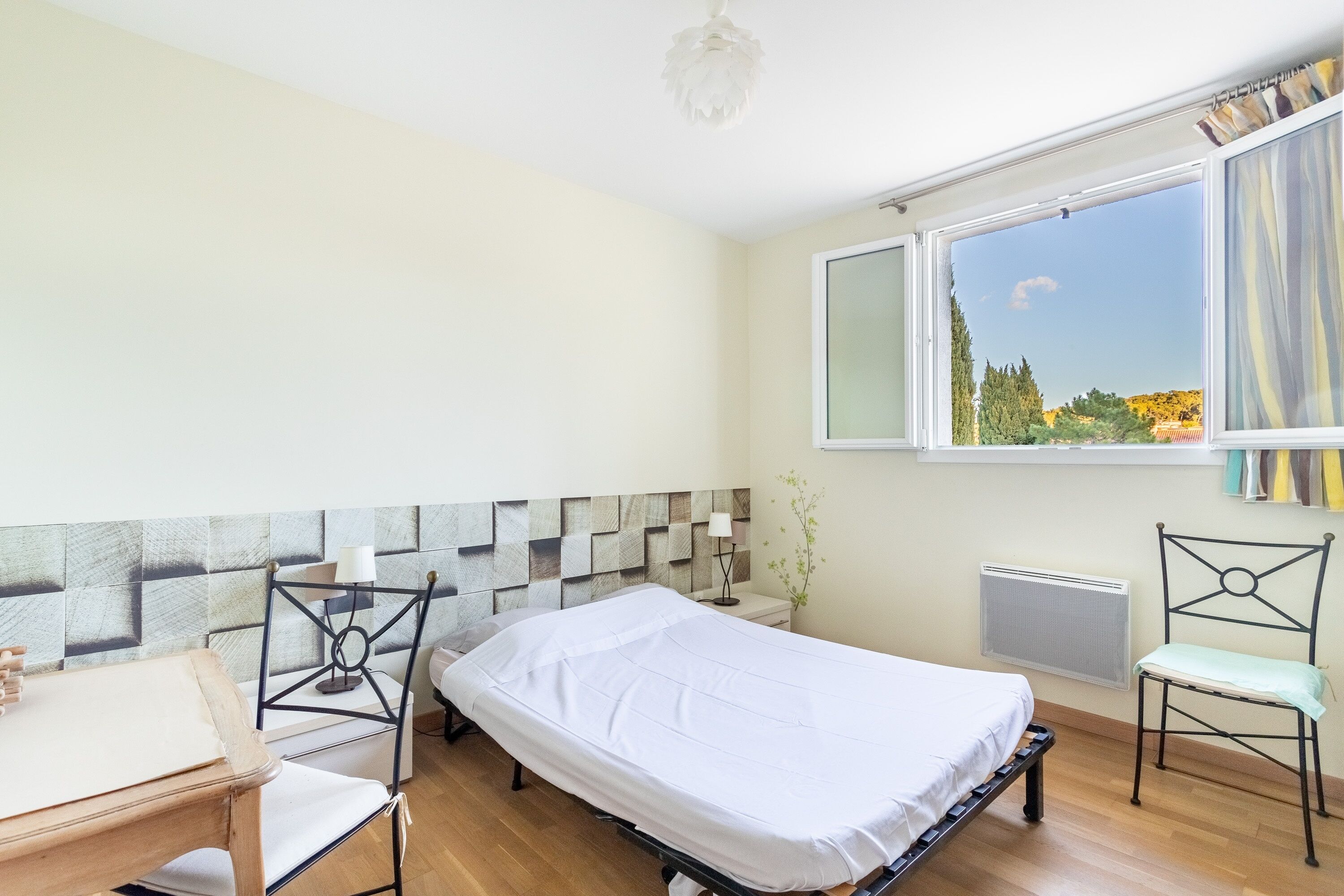 A bedroom with a bed, a desk, a chair, a radiator, and a window with a view of trees and a clear sky.