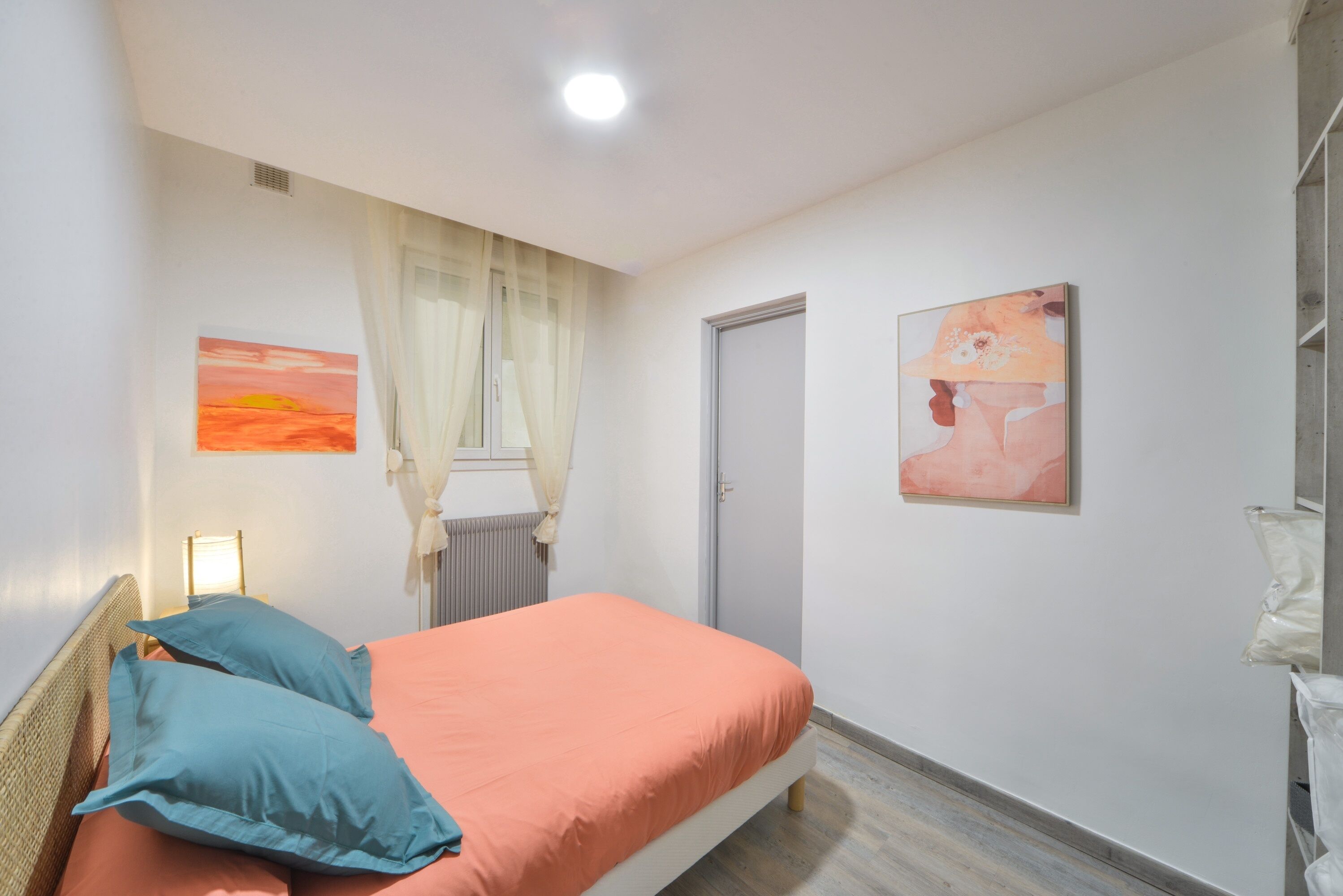 Un dormitorio con una cama, dos almohadas, un radiador, una puerta y dos cuadros en la pared.