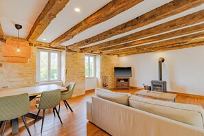 Ein Zimmer mit freiliegenden Holzbalken, einem Esstisch mit Stühlen, einer Couch, einem Fernseher und einem Kaminofen.