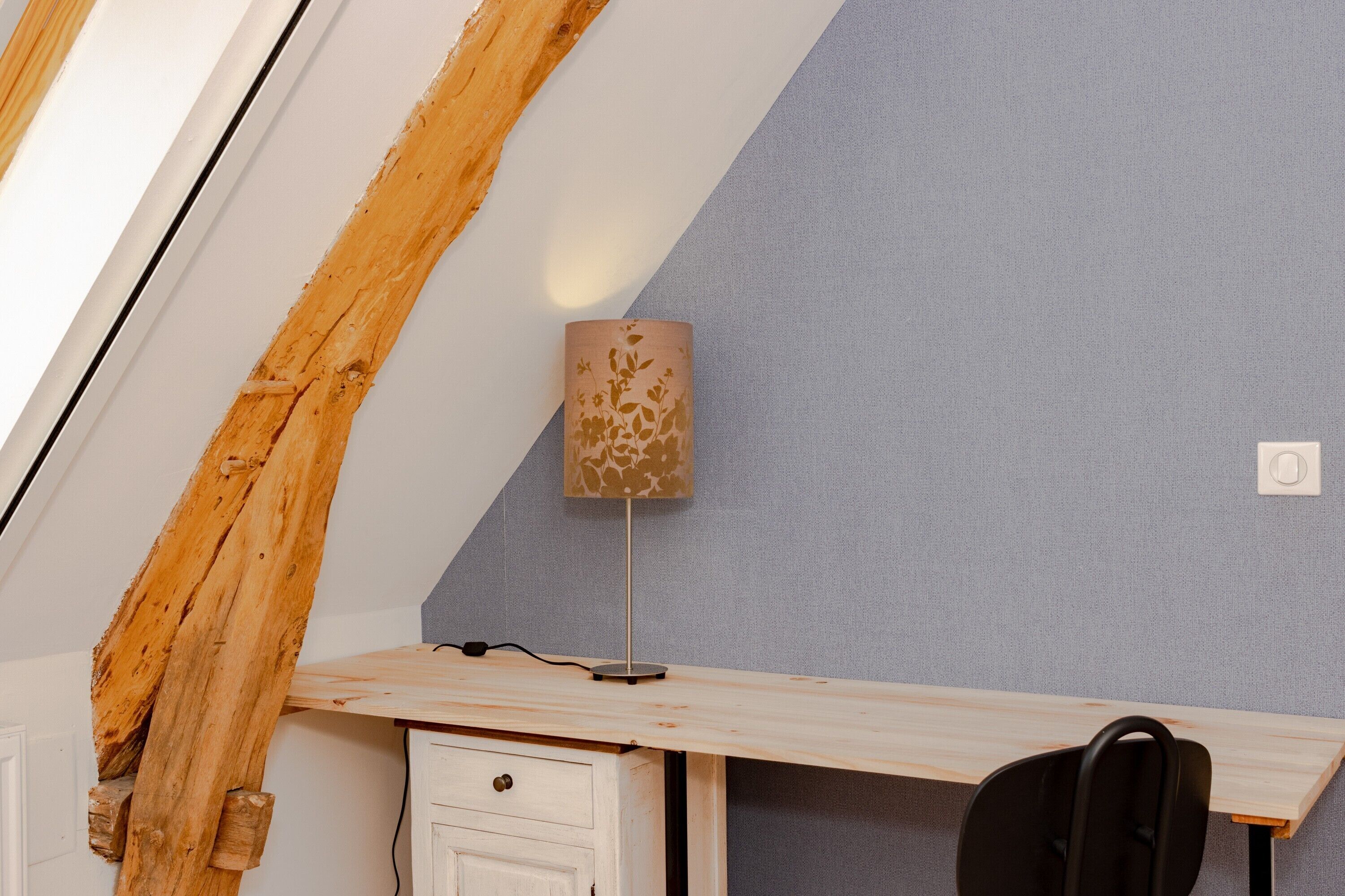 Een kamer met een schuine kap, een bureau met lamp, een stoel en een klein kastje.