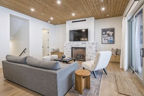 Uma sala de estar moderna com um sofá cinzento, uma poltrona branca, uma mesinha de madeira e uma lareira.