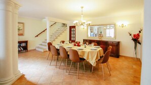 Zona pranzo con un tavolo rotondo apparecchiato per sei persone, circondato da sedie. Si nota una scala che conduce a un altro livello, una libreria e uno specchio appeso al muro.