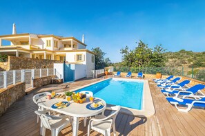 A poolside area with a dining table set for two, a pool with lounge chairs, and a building with a stone wall and a white fence.