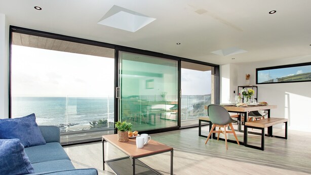 A modern living room with a large sliding glass door leading to a balcony with a view of the ocean. There is a sofa, a coffee table, and a dining area with chairs. - 4 bedroom accommodation in Torpoint (Torpoint)
