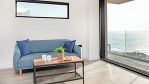 A modern living room with a blue sofa, a small wooden table with a plant and a plate of muffins, and a large window offering a view of the city. - 4 bedroom accommodation in Torpoint (Torpoint)