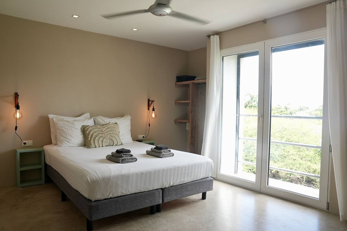 Un dormitorio con cama, lámparas de noche, ventilador de techo, ventana con cortinas y vistas al exterior con vegetación.