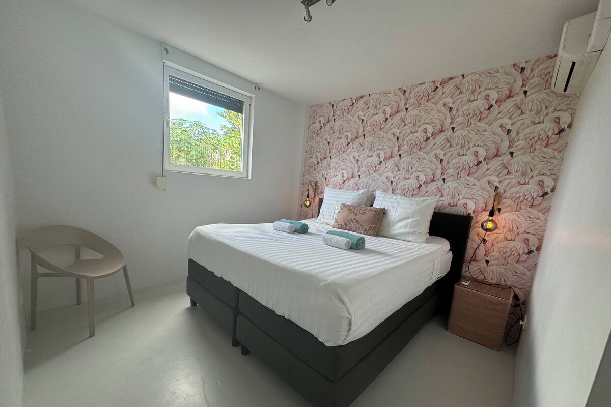 Un dormitorio con cama, ventana, silla y pared con estampado de flamenco.