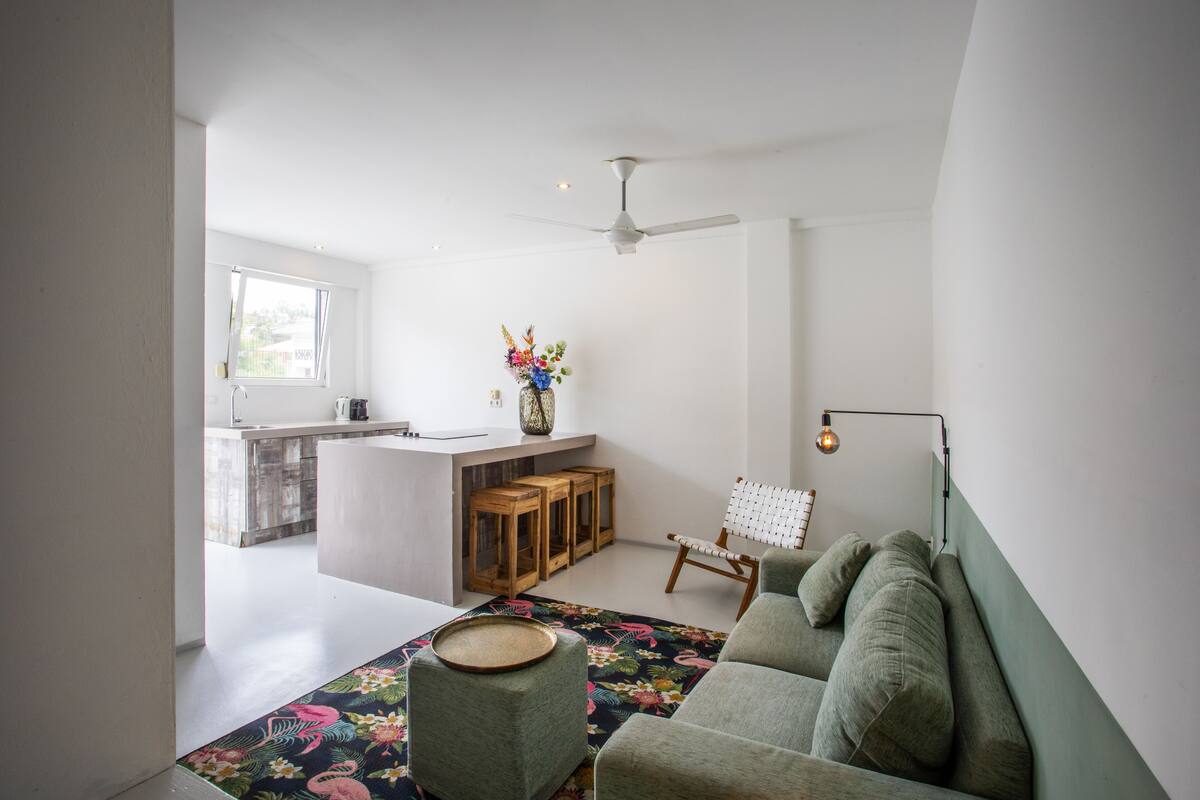 Una sala de estar moderna con un sofá, una mesa de centro y una pequeña mesa auxiliar de madera. Se ve una ventana que deja entrar luz natural y ofrece vistas al exterior.