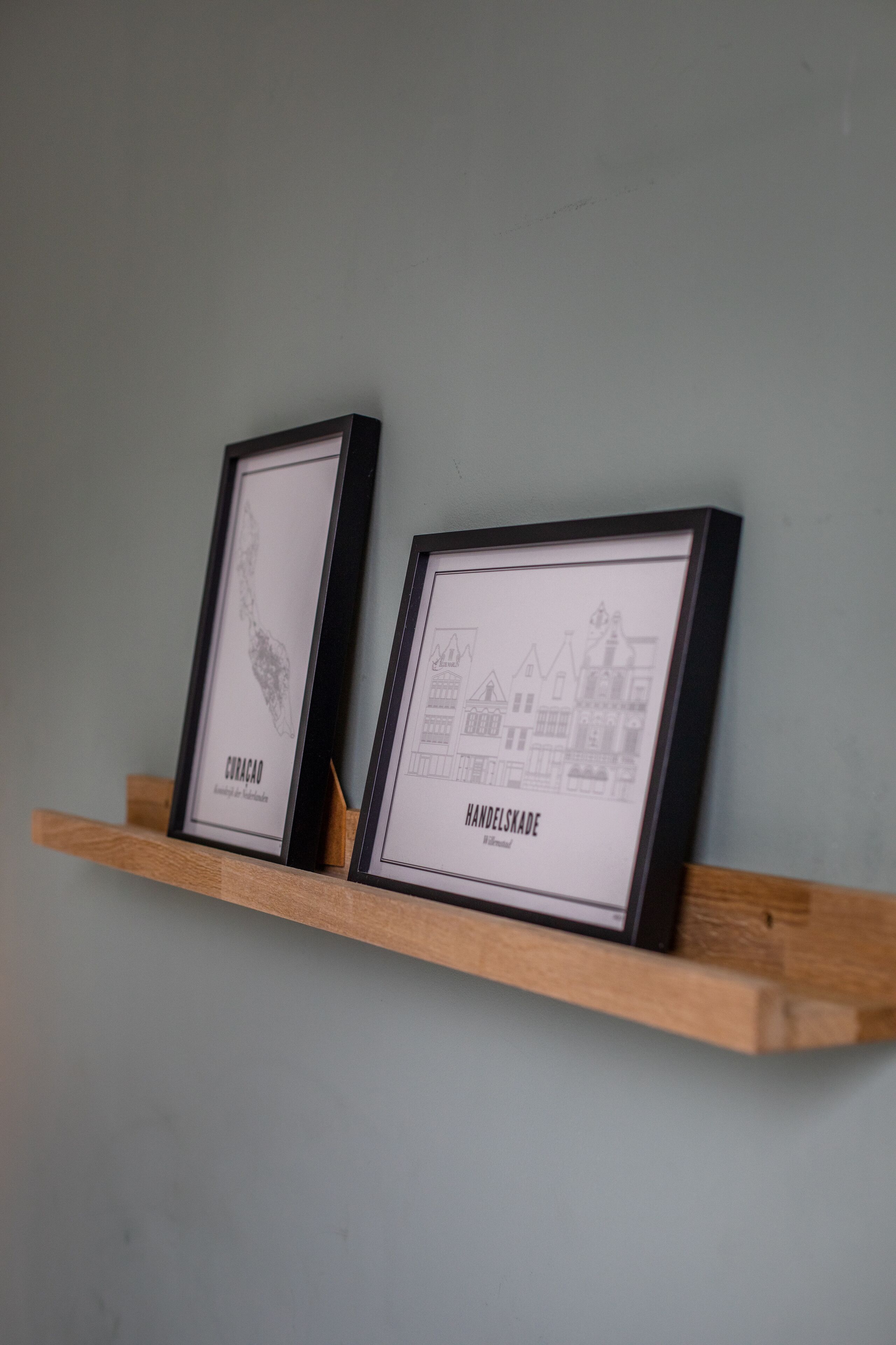 Dos dibujos arquitectónicos en blanco y negro enmarcados sobre una estantería de madera frente a una pared lisa.