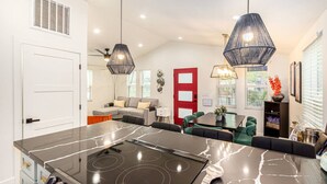 Een moderne keuken met een groot eiland, zwarte aanrechtbladen en een ingebouwde kookplaat. Er is een eethoek met een tafel en stoelen, en een woonkamer met een bank en een rode deur.