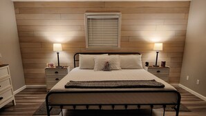 A bedroom with a bed, two bedside tables, a nightstand, a window with blinds, and a wall-mounted lamp. - Pickwick- Screened Patio ON Tennessee River 7th Nt FREE- Dogs Allowed

 (Morris Chapel)