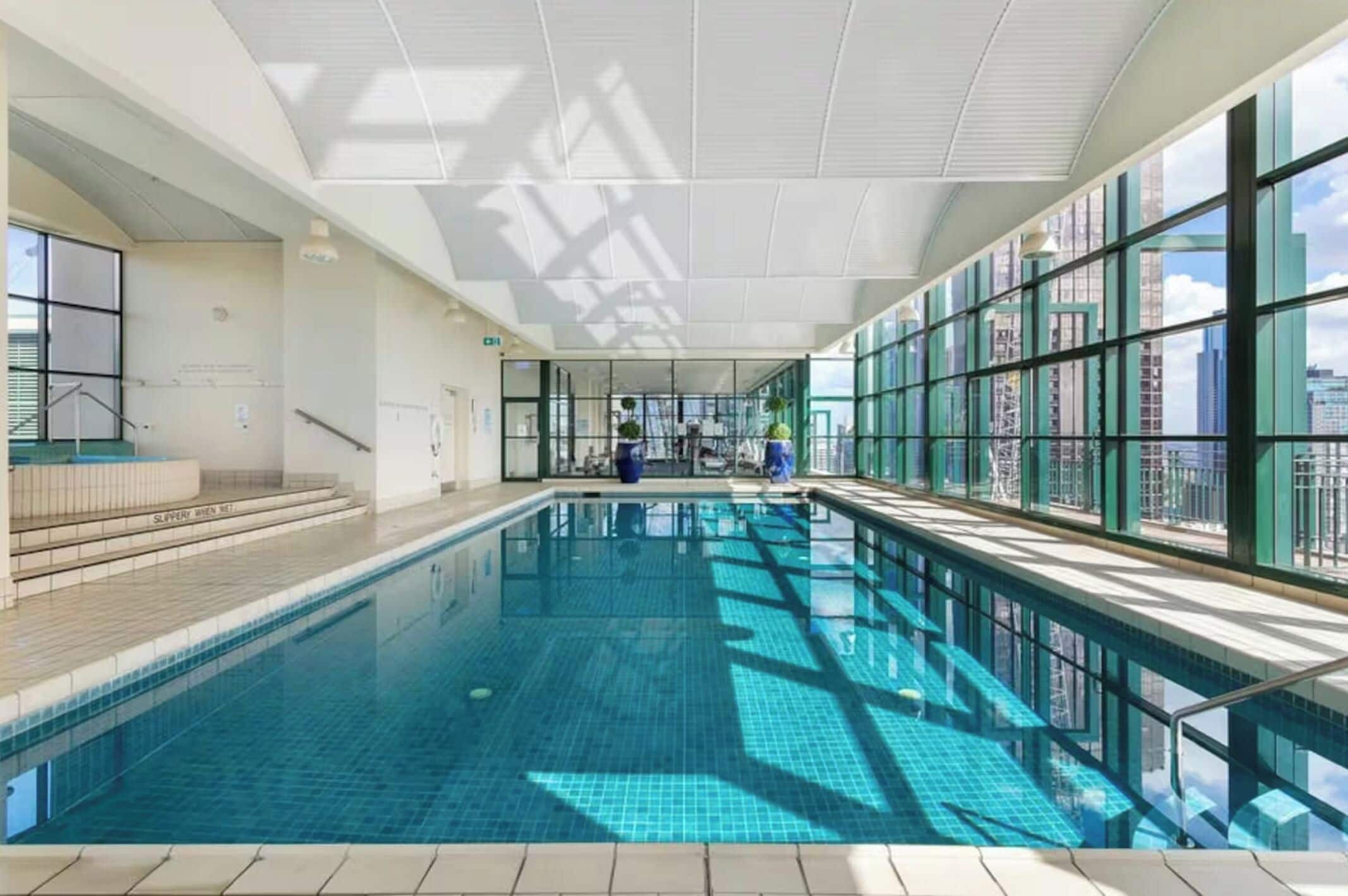 An indoor swimming pool with clear glass walls, a tiled floor, and a city view.