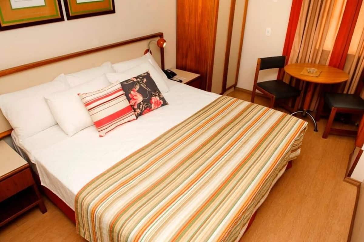A hotel room with a bed, a bedside table, a chair, a small round table, and curtains.