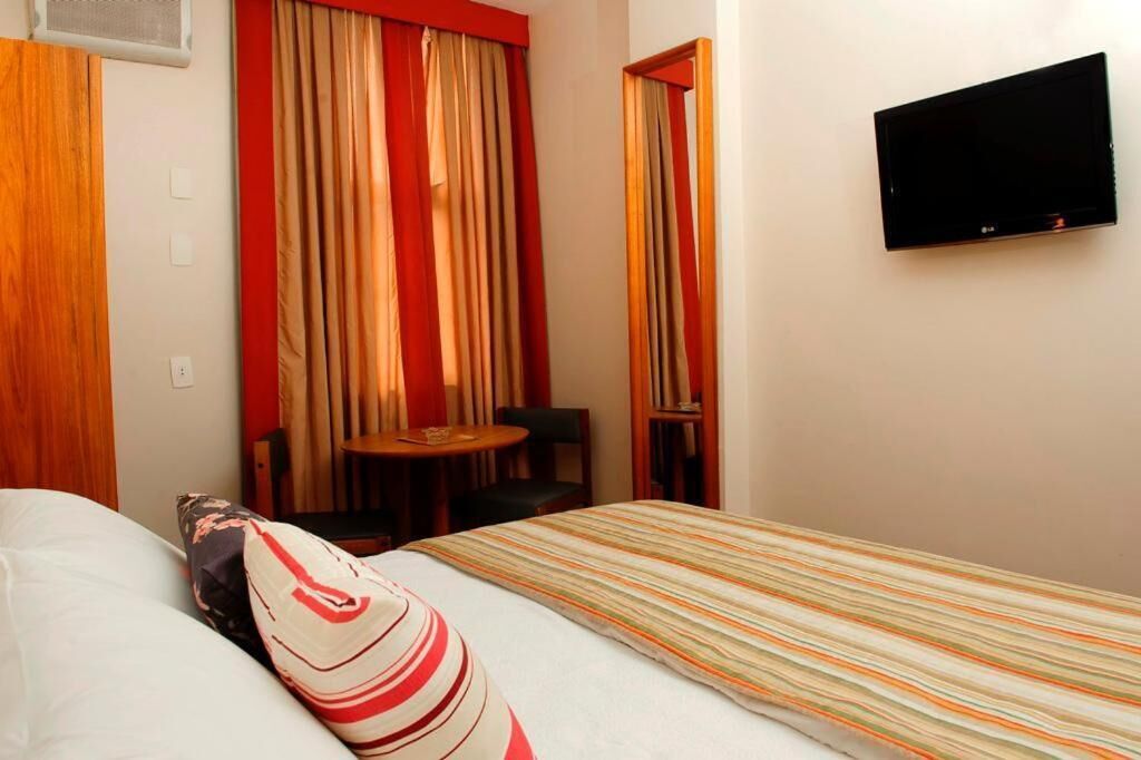 A hotel room with a bed, a TV mounted on the wall, a small table with a chair, and a window with curtains.