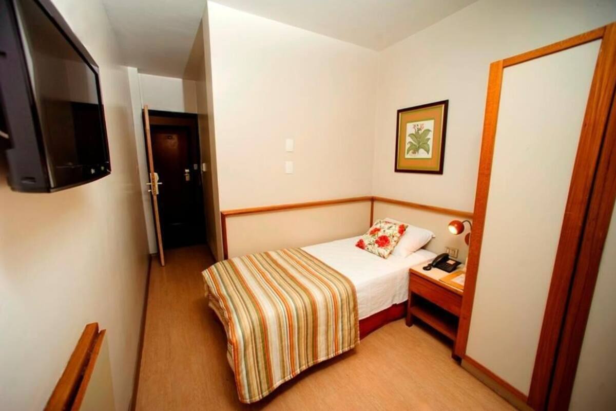 A small, narrow room with a single bed, a bedside table with a lamp, and a framed picture on the wall.