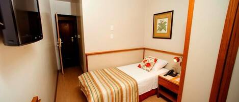 Una habitación pequeña y estrecha con una cama individual, una mesita de noche con lámpara y un cuadro en la pared.