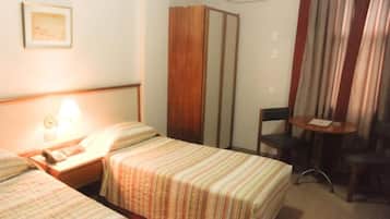 Quarto de hotel com duas camas, uma escrivaninha com luminária, uma cadeira e uma mesinha.