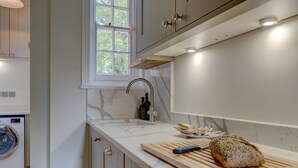 A modern kitchen with a built-in washing machine, a wooden cutting board with a loaf of bread, and a window with a view of trees. - Oxford Garden View | Oxford Holiday Let | 2 Beds & Sleeps 4 (Oxford)
