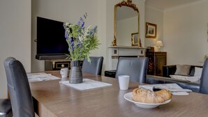 A dining area with a wooden table, chairs, a vase with flowers, a plate of pastries, a television, and a fireplace. - Oxford Garden View | Oxford Holiday Let | 2 Beds & Sleeps 4 (Oxford)
