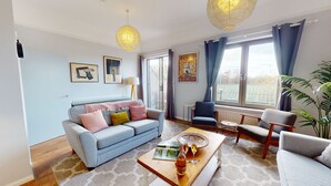 A modern living room with a grey sofa, a coffee table with a bottle and glass, and a large window with curtains. - Bannister House | Oxford Holiday Let | 5 Beds & Sleeps 9 (Oxford)