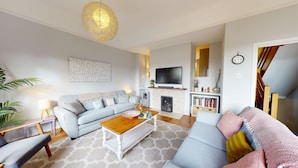 A living room with a sofa, coffee table, and a fireplace. There is a television mounted above the fireplace and a bookshelf with books. - Bannister House | Oxford Holiday Let | 5 Beds & Sleeps 9 (Oxford)