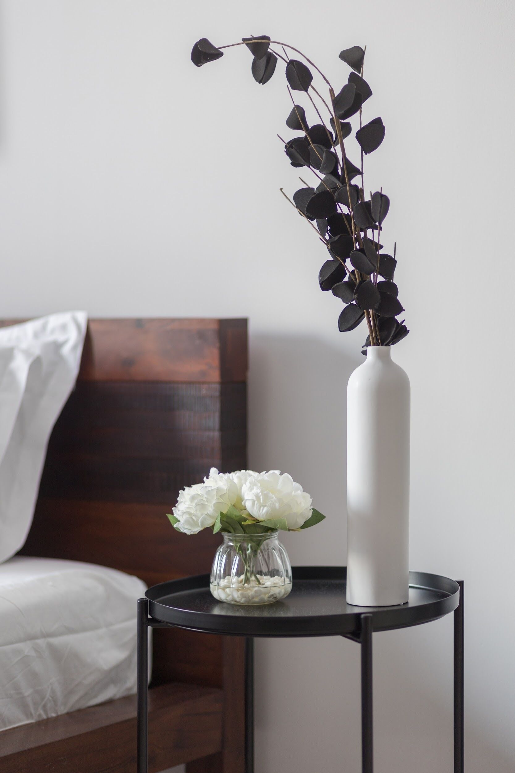 Ein Bett mit weißer Bettwäsche, ein kleiner Nachttisch mit schwarzer Oberfläche, darauf eine weiße Vase und ein Glas mit weißen Blumen, sowie ein dunkles Holz-Kopfteil.