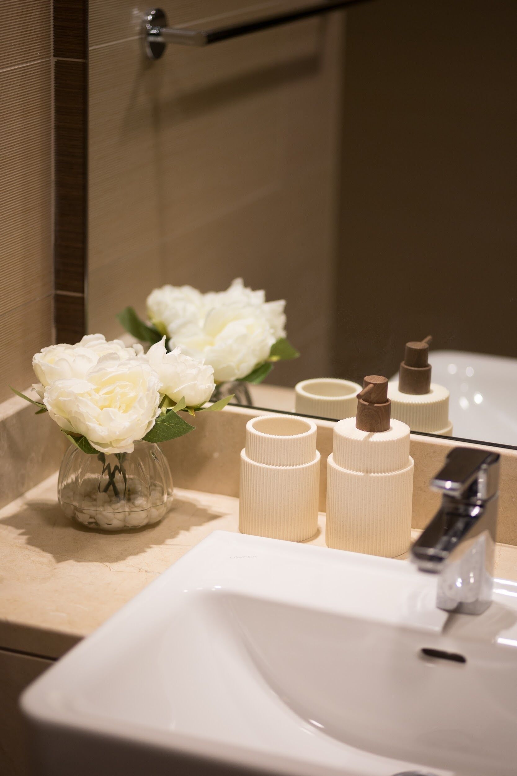 A bathroom sink with a mirror, a vase with white flowers, and three toiletry bottles.