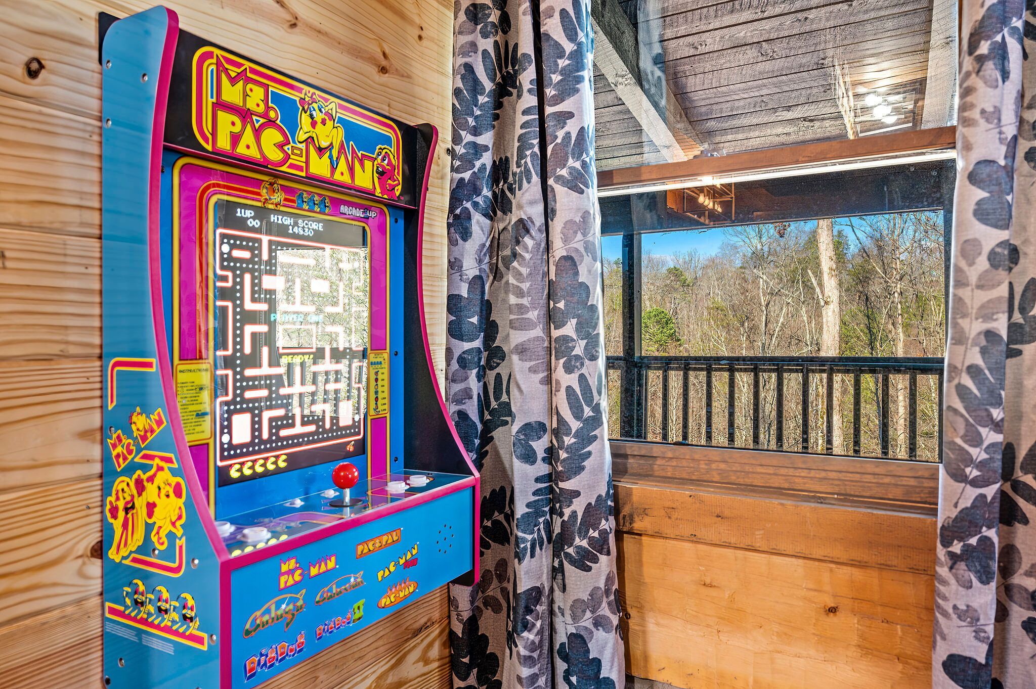 A Pac-Man arcade game in a wooden cabin with a view of trees through a window.