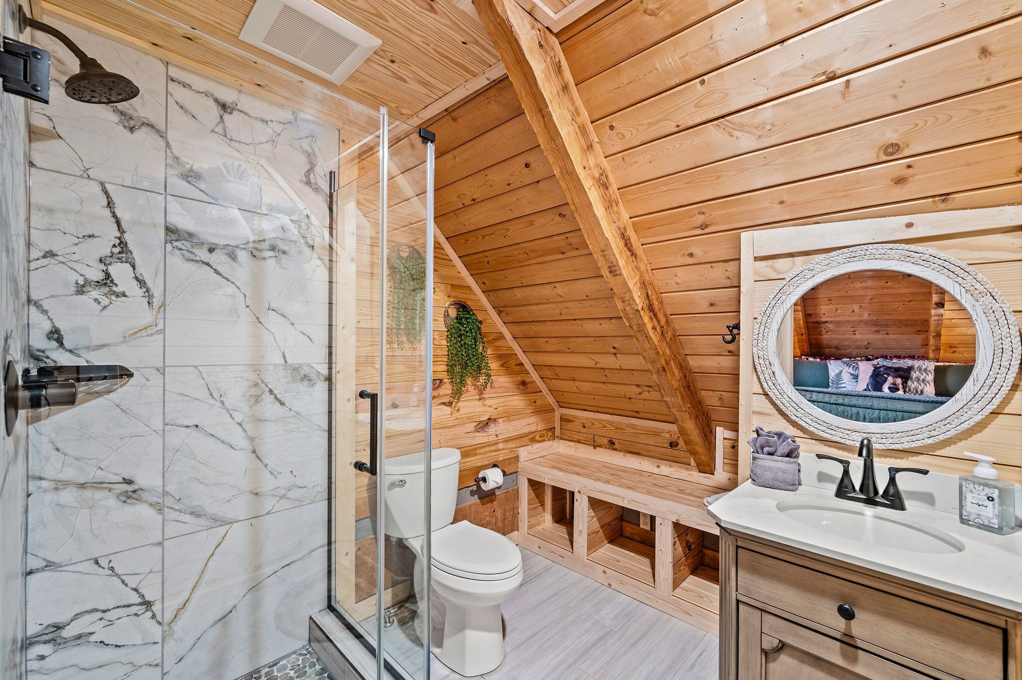 Un baño de cabaña de madera con cabina de ducha de vidrio, inodoro, lavabo con espejo y un banco.