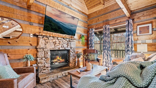A cozy log cabin interior with a stone fireplace, wooden walls, and a large window with curtains. - The Willows (Sevierville)