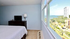 Un dormitorio con cama, cómoda, televisor y ventana con vistas a edificios y vegetación.
