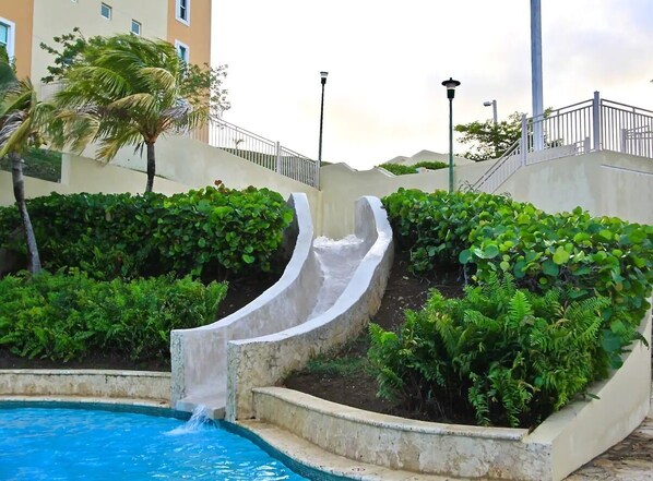 A swimming pool with a water slide, surrounded by lush greenery and a building in the background. - 608 Ocean & Marina Views 3 Bedroom 2 Bathroom Luxury Condo (Fajardo)