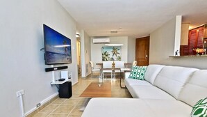 Una sala de estar moderna con un televisor de pantalla plana, un comedor con una mesa y sillas, y un sofá blanco con cojines estampados.