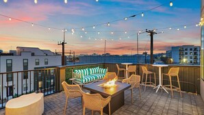 A rooftop terrace with string lights, a fire pit, outdoor furniture, and a view of the cityscape at dusk.
