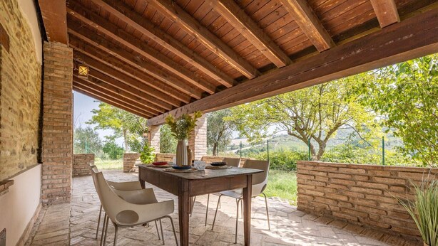 Een terras buiten met een houten tafel en stoelen, een bakstenen muur en uitzicht op bomen en heuvels.