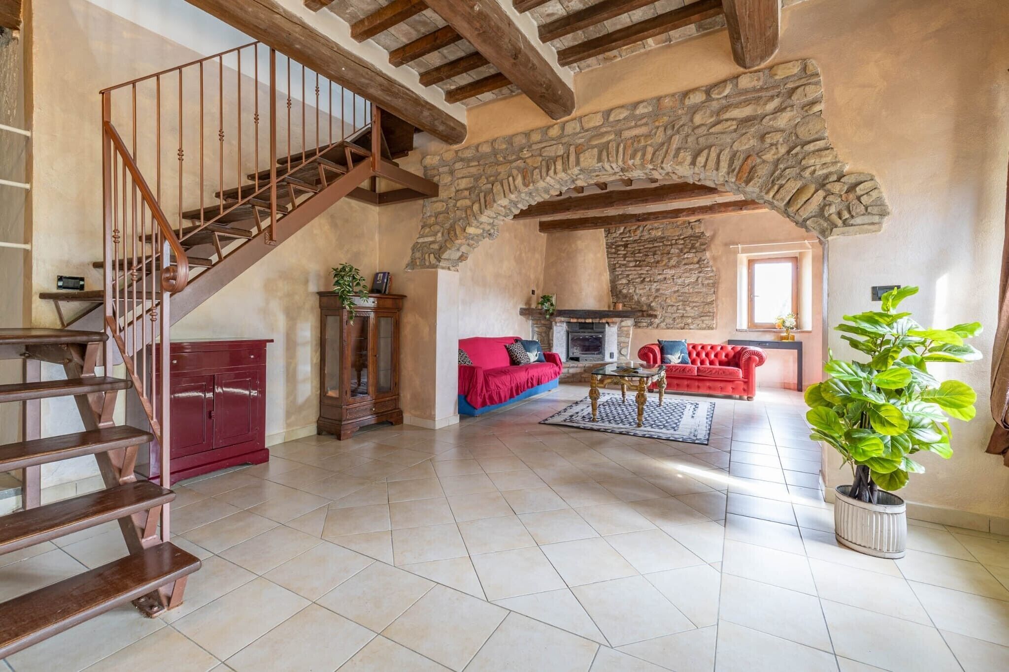 A spacious living area with a stone archway, a wooden staircase, a red sofa, a fireplace, and a potted plant.